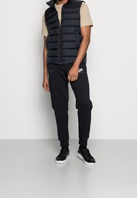 Svart vadderad väst med hög krage över en ljusbeige t-shirt, parat med svarta joggingsbyxor med Nike-logotyp. Svarta sneakers fulländar looken.