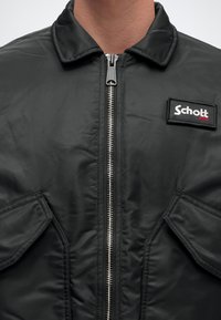 Veste noire zippée avec col, deux poches à rabat et un patch logo Schott NYC sur la poitrine.