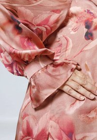 Fließende Blumenbluse aus zartem pinken Stoff, mit einem strukturierten Muster großer Blumen, lockeren Glockenärmeln und sanften Drapierungen.