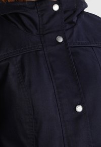 Granatowa kurtka z wysokim kołnierzem, z metalowymi zatrzaskami i przeszyciem w okolicy klatki piersiowej. Gładka tekstura materiału.