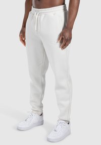 Weiße Fleece-Jogginghosen mit elastischem Bund und Bündchen, mit Seitentaschen und einem dezenten Logo auf der Vorderseite. Kombiniert mit weißen Turnschuhen.