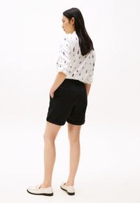 Pantaloni corti neri arrotolati realizzati in tessuto intrecciato, abbinati a una camicia bianca con un motivo di veliero. Il modello è in piedi con le mani nelle tasche.