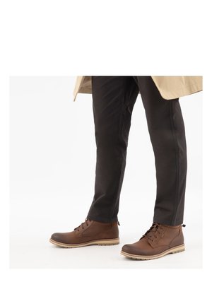 Beine mit schwarzer Hose und braunen Leder-Schnürstiefeletten mit beigen Sohlen auf weißem Hintergrund.