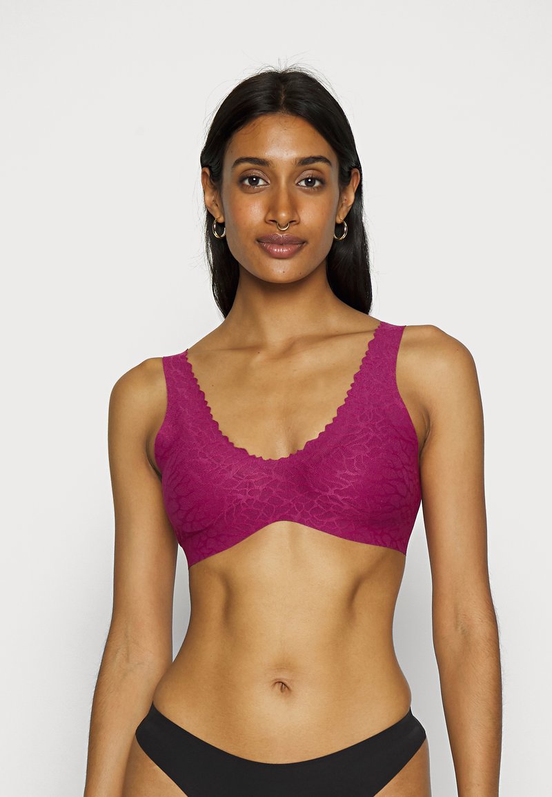 Kvinne iført en fuchsia blond bh og svart undertøy, står mot en enkel lys bakgrunn, vender mot kameraet med et nøytralt uttrykk.