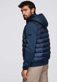 Gilet imbottito navy con cappuccio e orlo a costine. Caratterizzato da una texture liscia e un piccolo badge logo sulla spalla sinistra.