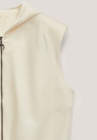 Sweat à capuche sans manches écru avec une fermeture éclair intégrale à l’avant et un tire-zip anneau en métal, présenté sur un fond clair.