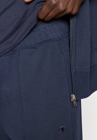 Χέρι που σηκώνει μπλε σκούρο φούτερ, δείχνοντας την τσέπη των αντίστοιχων μπλε σκούρων φόρμας και τη λεπτομέρεια ασημένιου φερμουάρ.