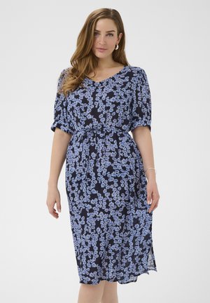 Žena v čiernych šatách po kolená s jemnou svetlofialovou kvetinovou potlačou, krátkymi nadýchanými rukávmi a opaskom v páse, stojaca pred jednoduchým pozadím.