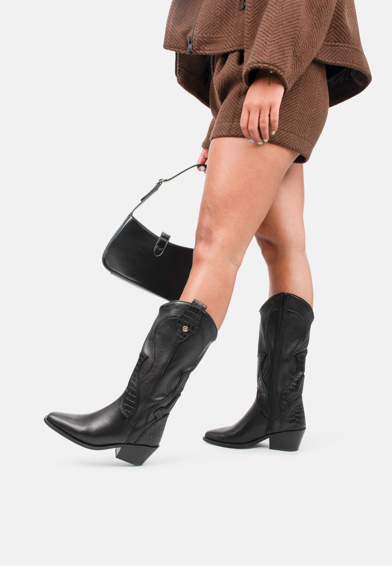 Las botas de vaquero negras hasta la rodilla cuentan con cuero texturizado, puntas puntiagudas y tacones de bloque. El diseño incluye costuras decorativas y acentos.
