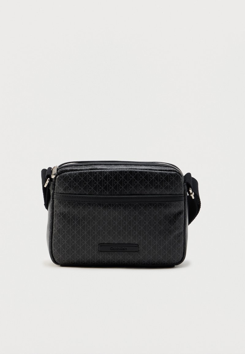 Μαύρη τσάντα χιαστί με γεωμετρικό σχέδιο, ομαλή υφή, κλείσιμο με φερμουάρ, λογότυπο ως διακοσμητικό και ρυθμιζόμενο λουρί. Συμπαγές ορθογώνιο σχήμα.