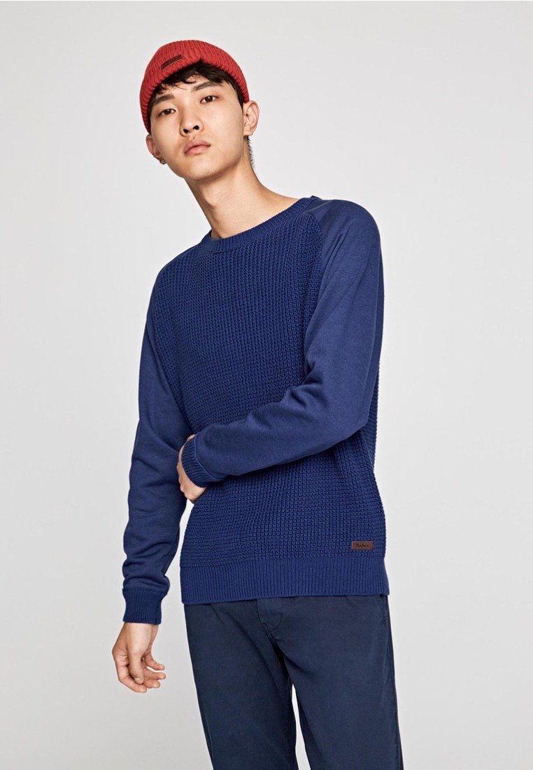 Pull en maille texturée bleu marine avec des manches longues, un col ras du cou et des poignets côtelés, associé à un pantalon sombre et un bonnet en tricot rouge.