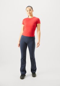 Rotes, tailliertes T-Shirt kombiniert mit dunkelblauen Sporthosen. Die Hosen haben einen breiten Bund und eine glatte Textur. Schwarzе Sportschuhe werden getragen.