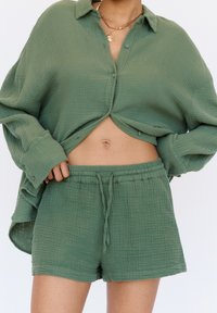 Zielony, teksturowany strój składający się z luźnej, zapinanej na guziki koszuli oraz dopasowanych szortów z elastycznym pasem i troczkiem, wykonany z lekkiego materiału.