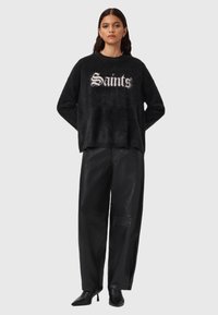 Musta pörröinen neule, jossa valkoinen "Saints"-teksti, yhdistetty korkeavyötäröisiin mustiin nahkahousuihin ja kärjellisiin mustiin korkokenkiin.