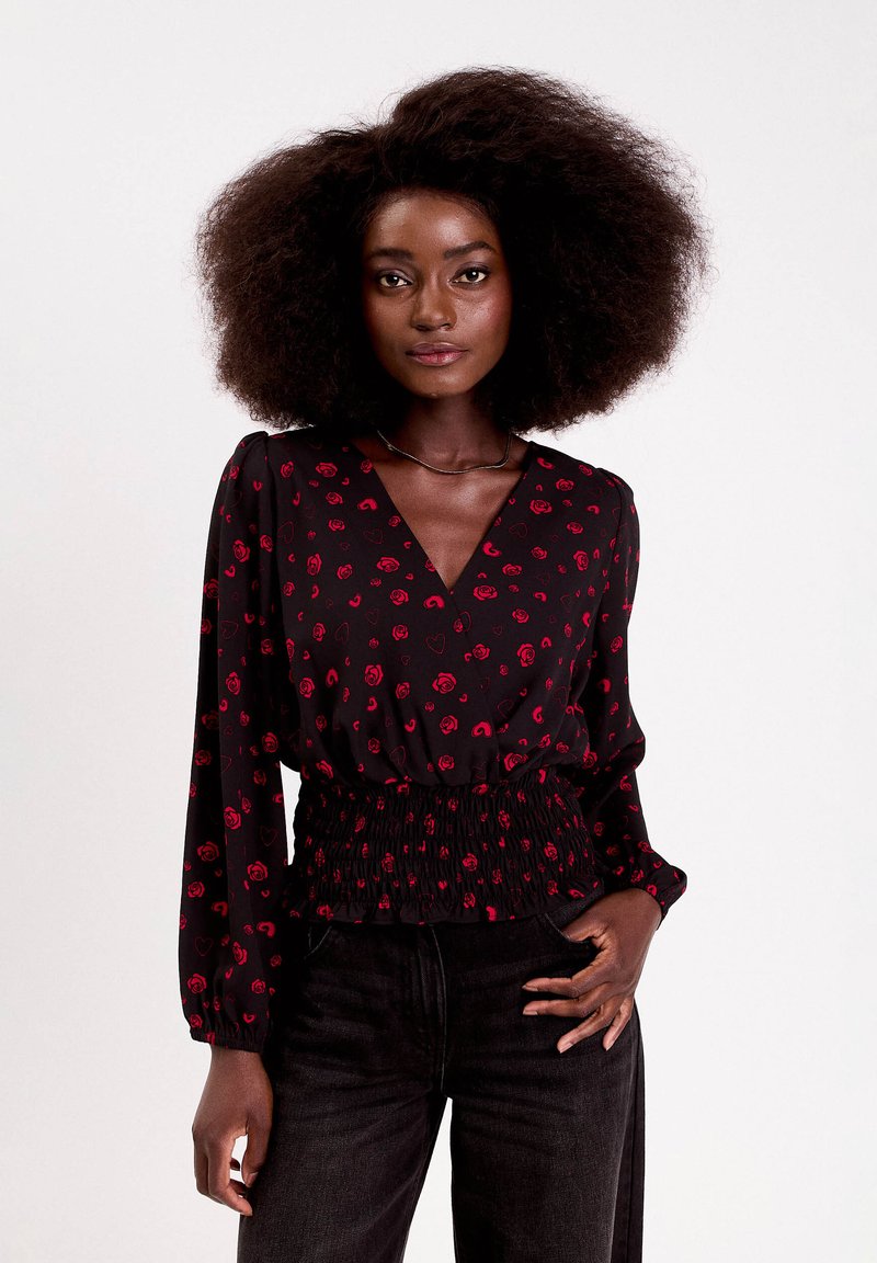 Czarna bluzka z czerwonym nadrukiem róż i serc, dekolt w serek, długie rękawy, marszczona elastyczna talia, zestawiona z ciemnymi dżinsami.