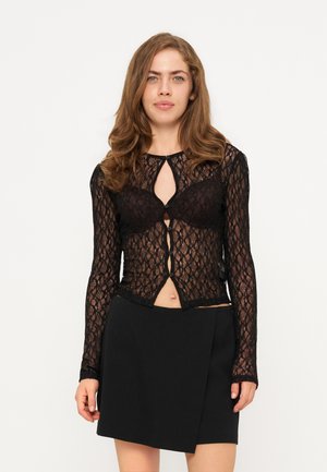Femme aux cheveux ondulés portant un haut noir en dentelle à manches longues boutonné sur un soutien-gorge noir et une jupe noire, debout devant un fond uni.