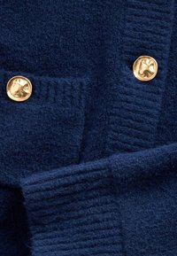 Tamsiai mėlynos spalvos tekstūruota medžiaga su banguotomis juostomis, puošta dviem apvaliais aukso spalvos mygtukais ir mažo kišenės elemento detale. Minkšta ir šilta išvaizda.