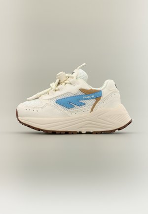 Krémszínű sportcipő vastag talppal, kék "Shadow" logóval, bézs díszítésekkel és húzózáras fűzőkkel, semleges háttéren.