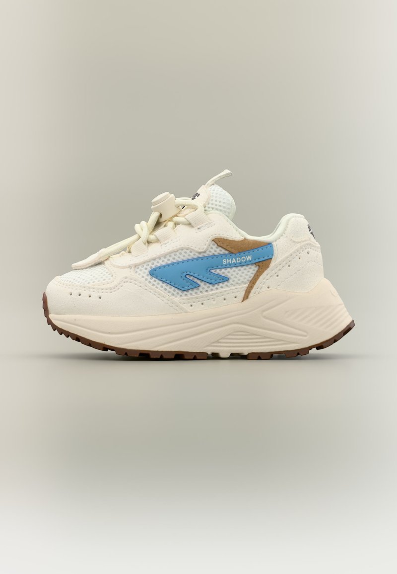 Krémszínű sportcipő vastag talppal, kék "Shadow" logóval, bézs díszítésekkel és húzózáras fűzőkkel, semleges háttéren.