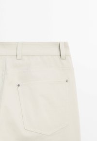 Vue arrière d’un pantalon beige clair montrant une ceinture avec passants et une poche arrière cousue avec rivets métalliques sur fond uni.