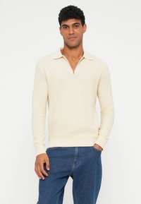 Uomo che indossa un maglione a polo con maniche lunghe e zip di colore crema, abbinato a jeans in denim blu, in piedi davanti a uno sfondo bianco.