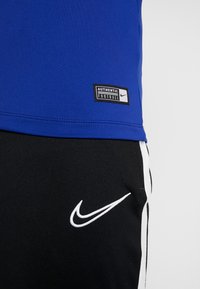 Sininen urheilupaita, jossa on musta "AUTHENTIC FOOTBALL" -merkki, ja mustat housut, joissa on valkoinen Nike-logo ja sivuraidat. Pehmeä kangas, istuva malli.