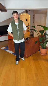 Žalia pūkinė liemenė dėvima ant baltos marškinėlių, juodi kelnės ir sidabrinės sportbačiai; stovintis kambaryje su medinėmis grindimis ir augalu.