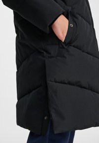 Sort puffet jakke med syede paneler, sidelommer og en trykknaplukning ved kanten. Stoffet har en glat, mat overflade.