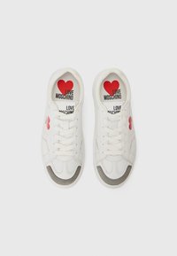 White sneakers with a red heart emblem on the side and a printed insole. Features flat laces, gray toe accents, and smooth leather texture.
