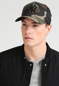 Tarnmuster-Baseballmütze mit schwarzem Mesh-Rücken, auf der Vorderseite mit einem auffälligen gestickten NY-Logo. Getragen mit einer schwarzen Bomberjacke und einem grauen Shirt.
