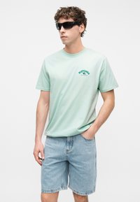 Νεαρός άνδρας φοράει μαύρα ηλιακά γυαλιά, μπλουζάκι Rip Curl σε μέντα πράσινο χρώμα και ανοιχτό μπλε τζιν σορτς, στέκεται με το ένα χέρι στην τσέπη.