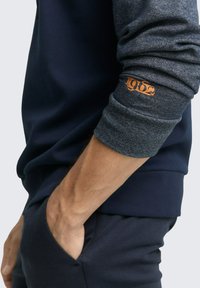 Granatowa bluza dresowa z szarymi teksturowanymi rękawami. Z nadrukiem "1962" na mankiecie, prążkowanym dołem oraz dopasowanym krojem.