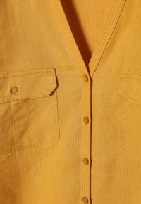 Mosterdgele shirt van linnenstof, met een V-hals, twee borstzakken en knoopsluitingen, met een soepele textuur.