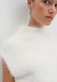 Weißer, gestrickter ärmelloser Pullover mit hohem Kragen, der eine weiche, flauschige Textur und leicht gerippten Stoff aufweist. Minimalistisches Design.