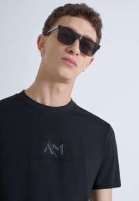 Camiseta negra con cuello redondo y detalles de logo en relieve. El modelo lleva gafas de sol oscuras, mostrando un diseño minimalista.