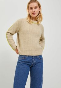 Maglione beige a righe con maniche lunghe, caratterizzato da accenti verde brillante lungo le maniche. Abbinato a jeans in denim blu.