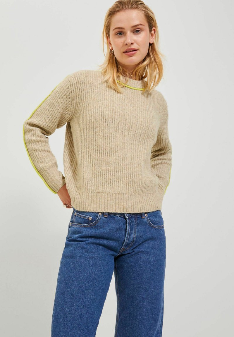 Maglione beige a righe con maniche lunghe, caratterizzato da accenti verde brillante lungo le maniche. Abbinato a jeans in denim blu.