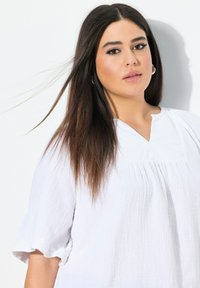 Blusa bianca e texturizzata con maniche corte a volant e scollatura a chiave. La modella ha i capelli lunghi, delicatamente illuminati su uno sfondo neutro.