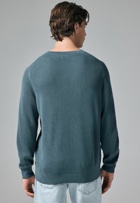 Pull en tricot texturé et côtelé en teal, avec des manches longues et un col rond, offrant une coupe décontractée et une couleur unie tout au long.