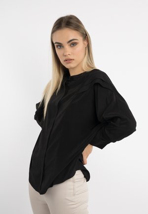 Młoda kobieta z prostymi blond włosami, ubrana w luźną czarną bluzkę i jasnobeżowe spodnie, stoi z jedną ręką na biodrze na białym tle.