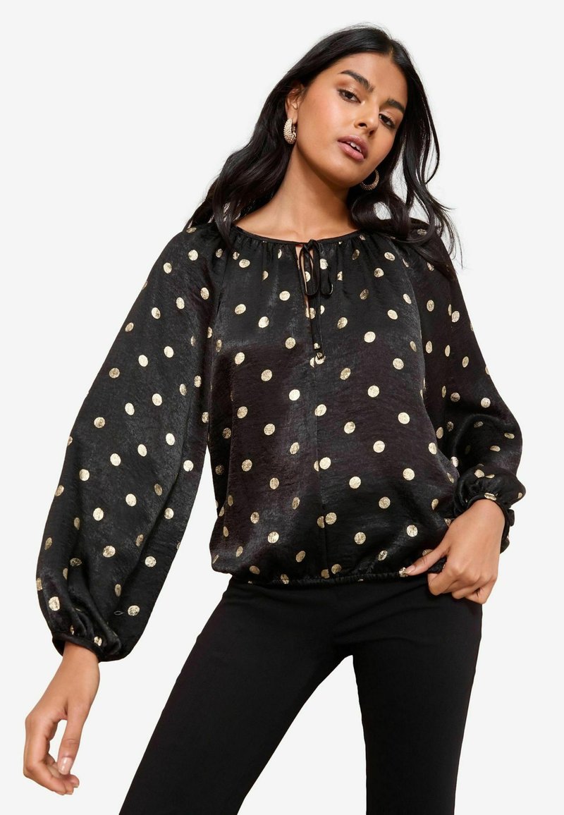 Musthave mustikas blouse, kus on kuldsed ringid, elastsed varrukad ja sidumisdetail kaelusel. Kanga pind on pehme ja kerge tekstuuriga.