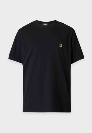 Jeune homme debout, une main dans la poche, portant un t-shirt noir avec un petit logo blanc, un pantalon noir et des baskets noires avec des semelles blanches.