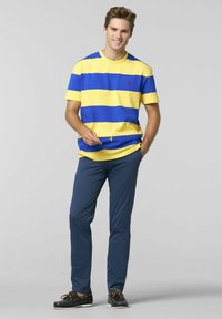 Gestreiftes T-Shirt in Blau und Gelb, Rundhals-Ausschnitt, kombiniert mit marineblauen Hosen und schwarzen Bootsschuhen. Auffälliges rotes Logo auf der Brust.