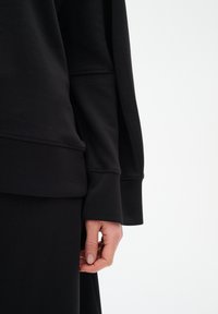 Felpa nera con maniche lunghe, caratterizzata da un taglio comodo e polsini a coste. Il materiale sembra morbido, con dettagli di cucitura visibili.