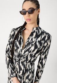 Schwarz- und cremefarbenes gemustertes Kleid mit knopflosem Ausschnitt, langen Ärmeln und Taillenband. Modell trägt braune Cateye-Sonnenbrille.