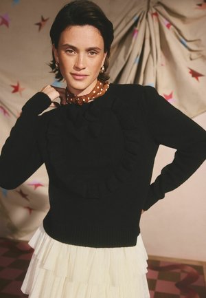 Schwarzer Pullover mit einem rüschendem herzförmigen Design, kombiniert mit einem braunen gepunkteten Kragen. Darunter ist ein mehrlagiger weißer Rüschensrock sichtbar.