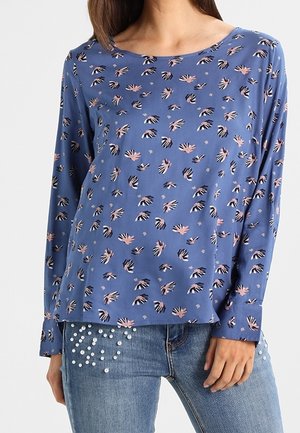 Frau trägt ein blaues Langarmoberteil mit pinken und schwarzen abstrakten Blumenmustern und blaue Jeans, verziert mit weißen Perlen.