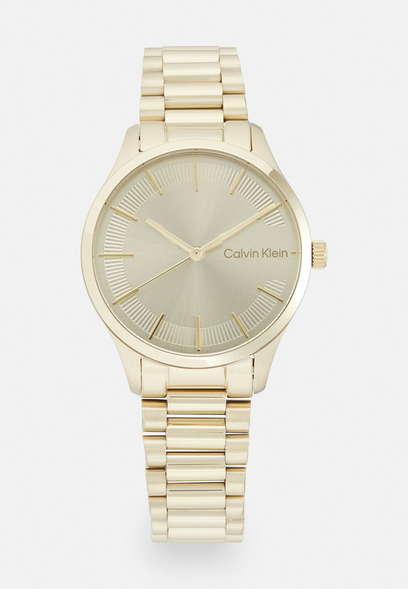 Montre dorée avec un cadran rond minimaliste, un cadran doré clair et un bracelet en maillon métallique. Dispose de repères horaires et de la marque Calvin Klein.