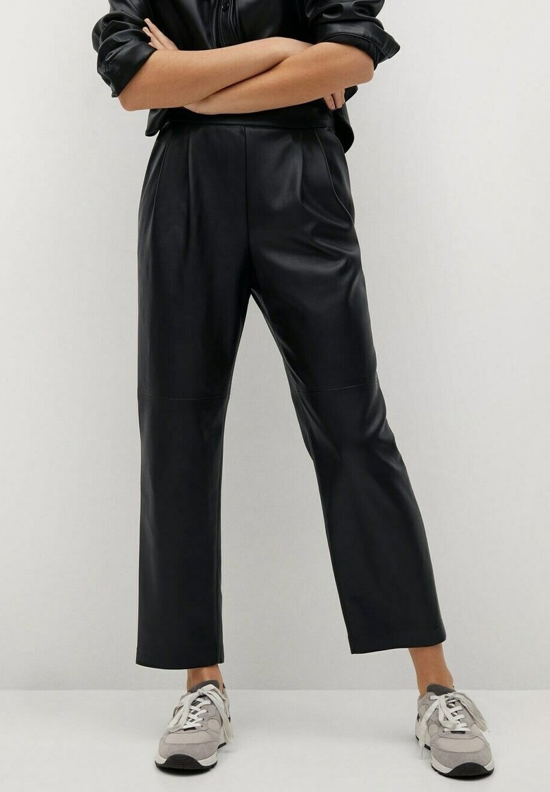 Pantalones de cuero sintético negros con corte recto, que presentan una textura suave y costuras en la parte frontal, combinados con zapatillas grises.