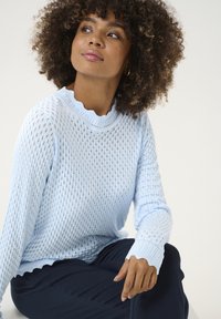 Hellblauer Strickpullover mit einem gewellten Ausschnitt und Bündchen, der ein offenes Strickmuster aufweist. Kombiniert mit dunkelblauen Hosen.